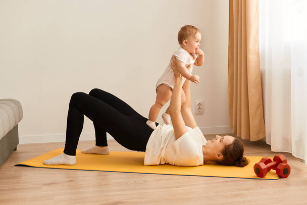 Side view portrait of young sporty mother doing physical yoga or pilates, exercises together with her baby daughter in her living room. Fitness, happy maternity and healthy lifestyle. - Foto, imagen