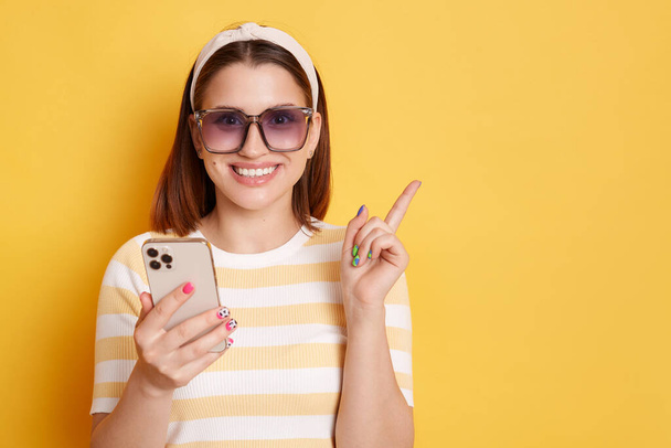 Portrait of happy smiling woman wearing sunglasses, striped T-shirt and hair band, using mobile phone and pointing finger at advertisement area, posing isolated over yellow background. - Photo, image