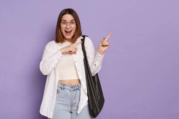 Portrait of cheerful woman with dark hair pointing fingers aside at copy space, demonstrating promo logo or company banner, wearing casual attire, posing isolated over purple background. - Fotoğraf, Görsel