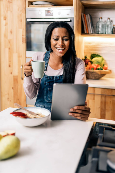Happy black woman drinking coffee in the kitchen at home and using tablet. - Fotografie, Obrázek