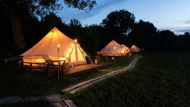 Палатки с горящими факелами, лампами и деревянными стульями при глампинге, лес вокруг, сумерки - Фото, изображение