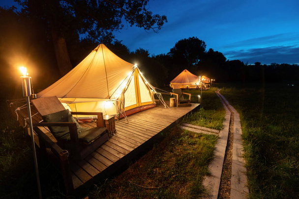 Tents with burning torches, lamps and wooden chairs at glamping, forest around, night - 写真・画像