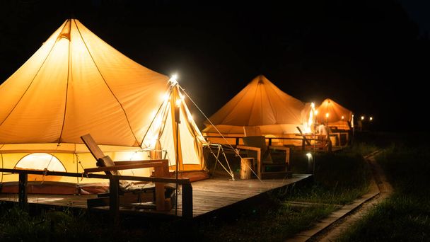 Палатки с горящими факелами, лампами и деревянными стульями при глампинге, ночь - Фото, изображение