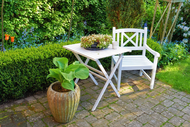 Patio krzesło i kwadratowy stół z dużą ilością roślin w ogrodzie. Meble ogrodowe do wypoczynku w lecie podczas ogrodnictwa kwiatowego. Rośliny doniczkowe na małym brukowanym dziedzińcu z zieloną trawą i drzewami. - Zdjęcie, obraz