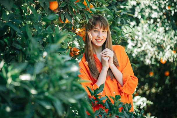 Happy young girl in orange dress is looking at camera by holding hands together on chest in orange garden  - Fotografie, Obrázek