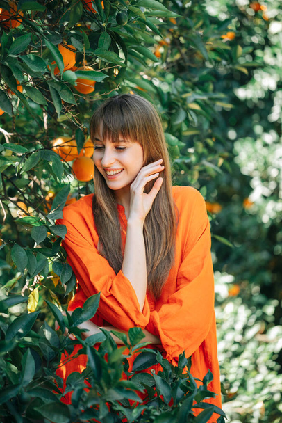 Happy young girl in orange dress is looking down by holding hand near face in orange garden  - Φωτογραφία, εικόνα