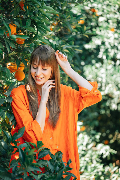 Happy girl in orange dress is posing to camera by looking down and holding up her hands in orange garden  - Photo, image