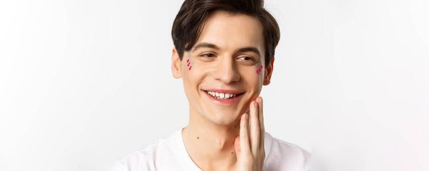 People, lgbtq and beauty concept. Headshot of beautiful gay man with glitter on face, smiling and looking happy, touching cheek after kiss, white background. - Foto, immagini
