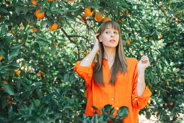 Молодая девушка в оранжевом платье смотрит в сторону, держа за руку возле уха в оранжевом саду  - Фото, изображение