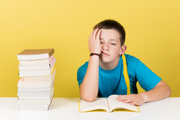 Демотивированный мальчик, читающий книгу на желтом фоне. Скучный студент держит голову с рукой рядом с грудой книг - Фото, изображение