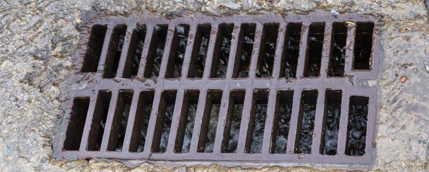 Cast iron grid of the storm drain mounted in a concrete surface with flowing down rainwater and small debris during a rain, panoramic view close-up - Foto, Imagem
