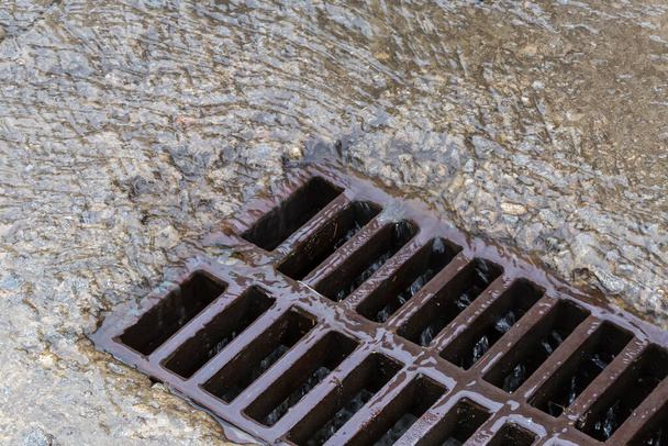 Fragmento de la rejilla de hierro fundido del desagüe de tormenta montado en una superficie de hormigón y que fluye por el agua de lluvia con pequeños escombros durante una lluvia - Foto, Imagen