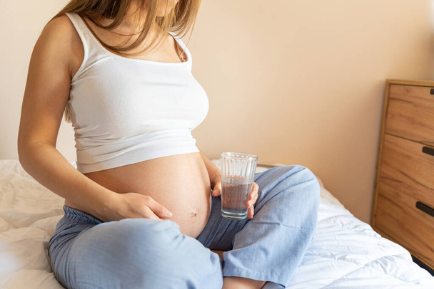 妊娠中の飲料水。きれいな妊娠飲料水。幸せな妊婦の水のガラスを保持する。妊娠、出産、医療の概念 - 写真・画像