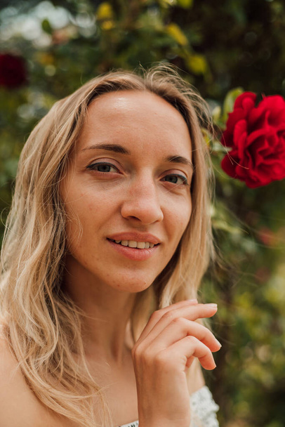 Portrait of a young tired woman near red roses. Behind beautiful nature. Next to a woman is a bush of red roses - Φωτογραφία, εικόνα