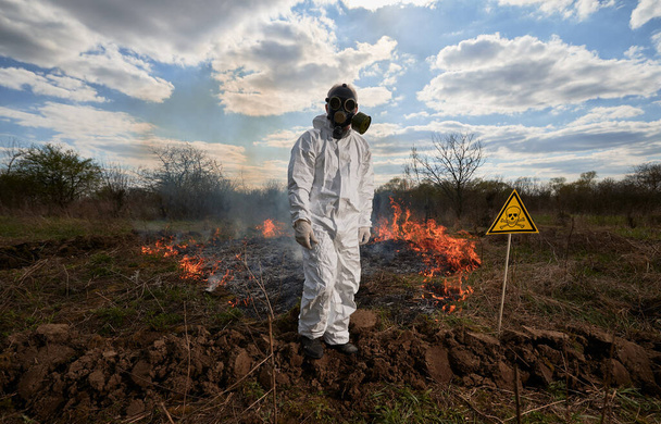 Fireman ecologist working in field with wildfire. Man in gas mask near burning grass with smoke and yellow triangle with skull and crossbones warning sign. Natural disaster concept. - Foto, immagini