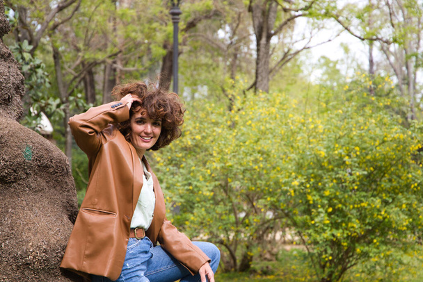 beautiful Spanish woman with curly hair is leaning on a tree trunk in the park. The woman is happy and enjoying her holiday in the city. Concept happiness, fashion and beauty. - Photo, Image