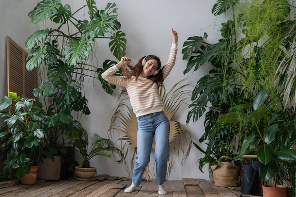 Carefree young woman with long hair in wireless headphones listening to music with pleasure, enjoying the moment, relaxing dancing on wooden floor in cozy home garden with monstera and tropical plants - Photo, Image