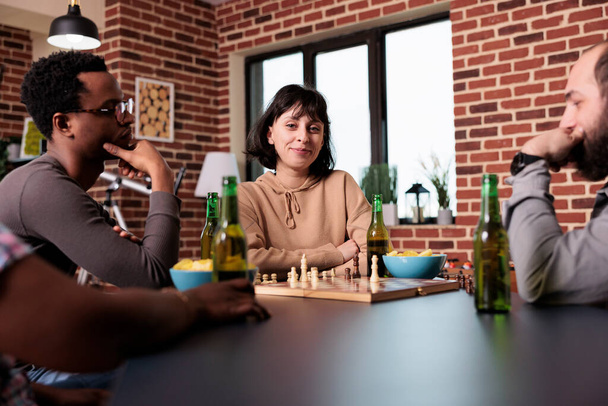 Radosna kobieta siedząca z przyjaciółmi w salonie i grająca w szachy. Szczęśliwi zróżnicowani ludzie cieszący się strategicznymi grami planszowymi, jednocześnie relaksując i spożywając przekąski i napoje. - Zdjęcie, obraz