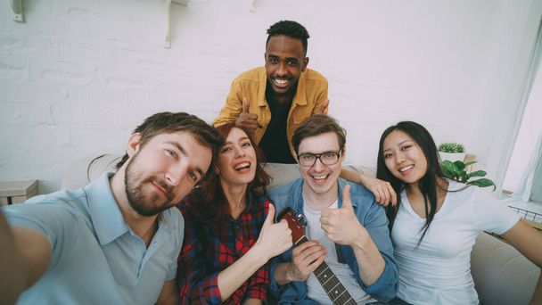 Point of view shot of multi ethnic group of cheerful friends taking selfie photos on smartphone camera while celebrating at party at home - 写真・画像
