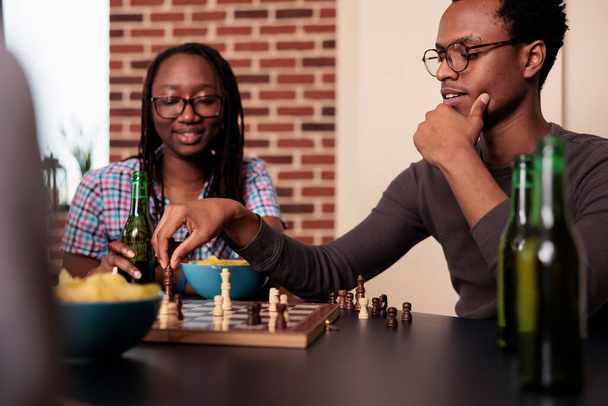 Зачаровані афроамериканські друзі сидять за столом у вітальні, граючи в шахові ігри. Веселі розумні люди вдома відпочивають з грою в шахи, насолоджуючись закусками та напоями.. - Фото, зображення
