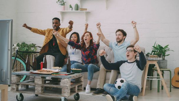 Group of happy friends watching sports game on TV at home indoors. They are happy about their favorite team win competition - 写真・画像