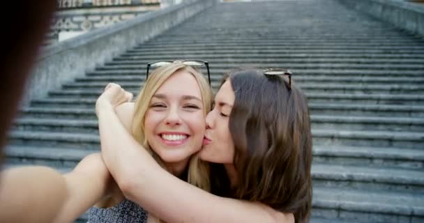 Uma mulher alegre beijando seu amigo na bochecha. Uma jovem mulher tirando uma selfie enquanto seu amigo a abraça carinhosamente. - Filmagem, Vídeo