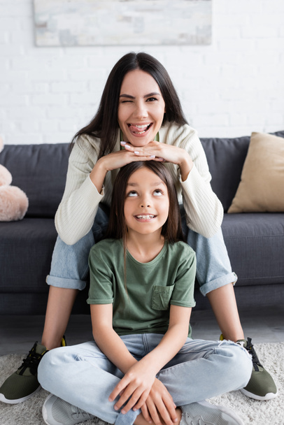 happy brunette babysitter sticking out tongue and sitting on couch near cheerful child - Foto, imagen