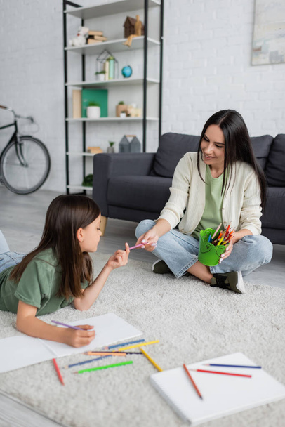 cheerful nanny passing pencil to girl lying on carpet near blank paper  - Photo, Image