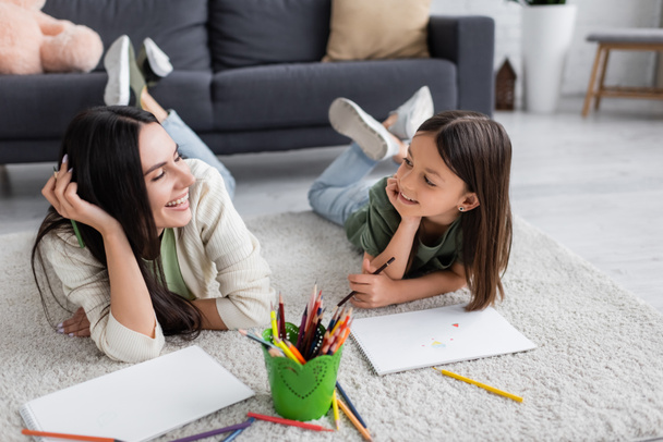 smiling nanny and girl looking at each other while lying on carpet near blank papers in living room  - Foto, Bild