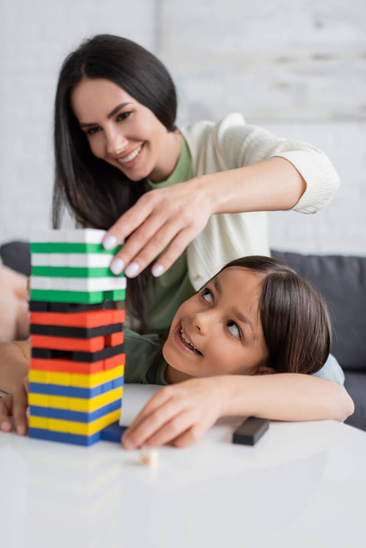 cheerful babysitter playing wood blocks game with pleased girl in living room - Foto, Imagen