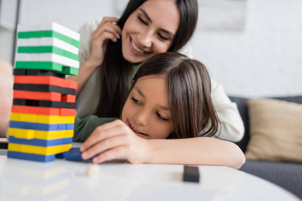 happy babysitter playing wooden tower game with pleased girl in living room - Foto, Bild
