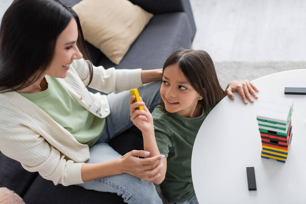 top view of happy babysitter playing wooden tower game with positive girl in living room - Foto, afbeelding