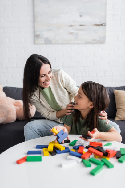 cheerful babysitter looking at girl near multicolored wooden blocks on table - Photo, Image