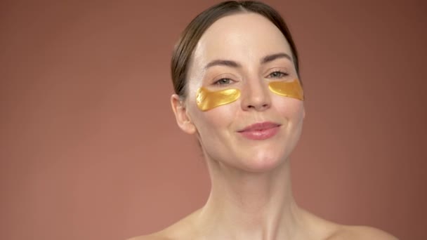 Bare shoulders beautiful female with eye patches touching face and looking at camera during skin care routine against brown background - Séquence, vidéo
