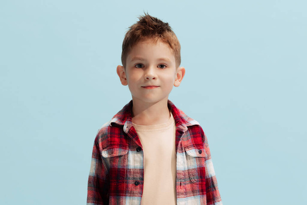 Portrait of little boy, child in checkered shirt looking at camera, barely smiling, posing isolated over light blue studio background. Concept of childhood, family, emotions, lifestyle, fashion, ad - Foto, Imagem