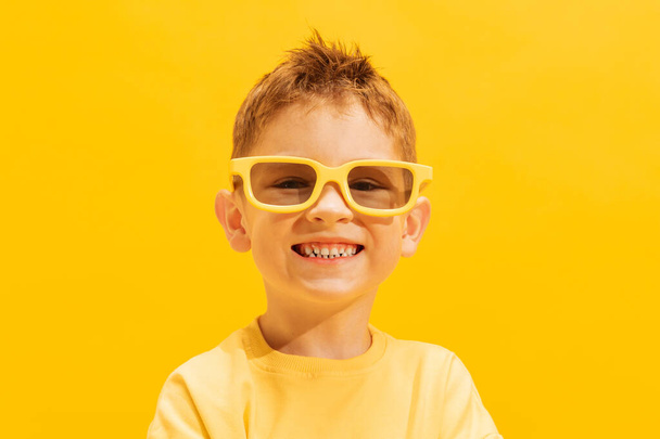 Porträt eines lächelnden kleinen Jungen, eines Kindes in hellem T-Shirt und Sonnenbrille, das isoliert vor gelbem Hintergrund posiert. Sommerurlaub. Konzept von Kindheit, Familie, Emotionen, Lebensstil, Mode, Werbung - Foto, Bild