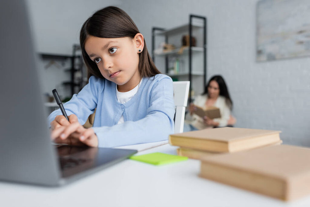 girl writing near blurred laptop while doing homework near nanny on background - Foto, immagini