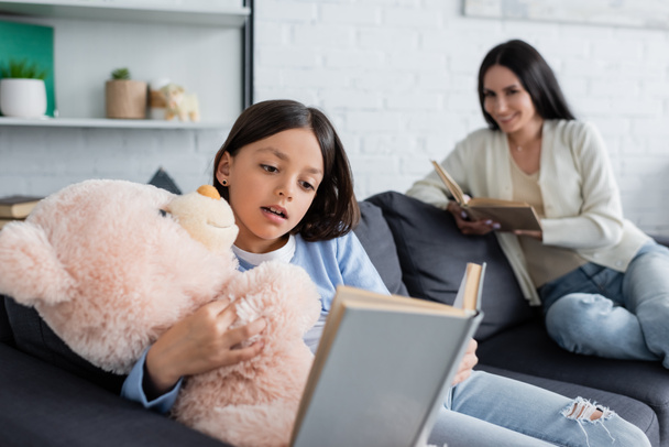 girl embracing teddy bear while reading book near nanny smiling on blurred background - Foto, afbeelding
