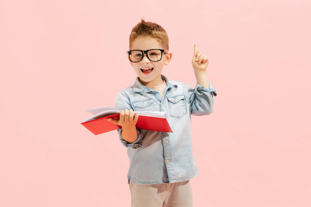 Portrait of little boy, child in jeans t-shirt and glasses, posing with book isolated over pink background. Education look. Concept of childhood, family, emotions, lifestyle, fashion, ad - Foto, immagini