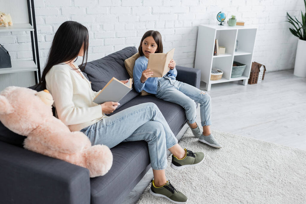 full length of child and nanny sitting with books on couch in living room - Foto, Imagem