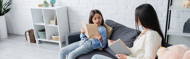 girl looking at babysitter while reading book on couch in living room, banner - Fotografie, Obrázek