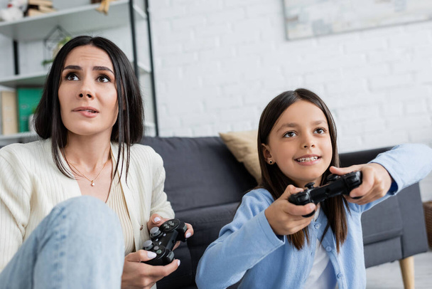 KYIV, UKRAINE - MAY 23, 2022: worried babysitter biting lip while gaming with cheerful girl at home - Fotografie, Obrázek