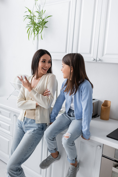 cheerful nanny talking to girl sitting on worktop in kitchen - Photo, image
