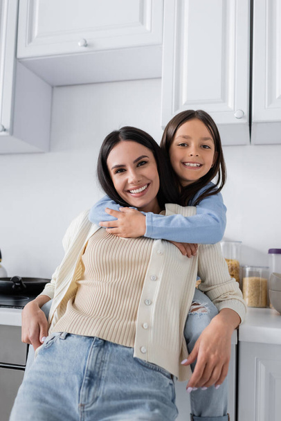 cheerful girl hugging happy nanny and smiling at camera in kitchen - Foto, Imagem