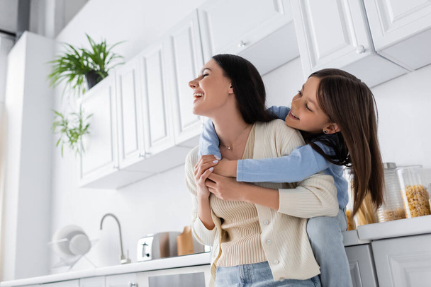 happy girl embracing laughing babysitter in kitchen - Photo, image