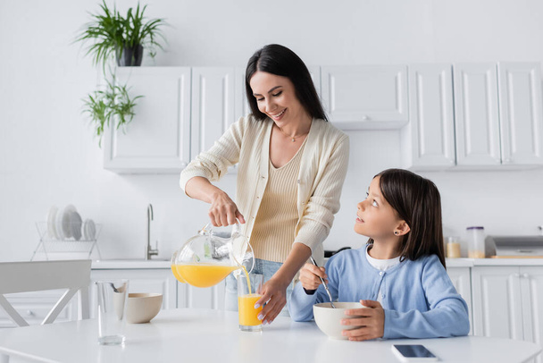 cheerful babysitter pouring orange juice near child having breakfast in kitchen - Foto, Imagem