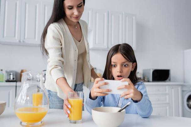 niesamowite dziewczyna patrząc na telefon komórkowy w pobliżu uśmiechnięta niania serwująca sok pomarańczowy na śniadanie - Zdjęcie, obraz