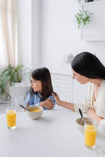 nanny calming upset girl looking away near corn flakes and orange juice in kitchen - Foto, afbeelding