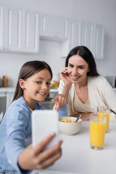 happy girl showing blurred mobile phone to babysitter during breakfast - Foto, imagen