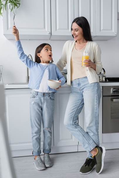 astonished girl holding spoon in raised hand near nanny with glass of orange juice - Zdjęcie, obraz